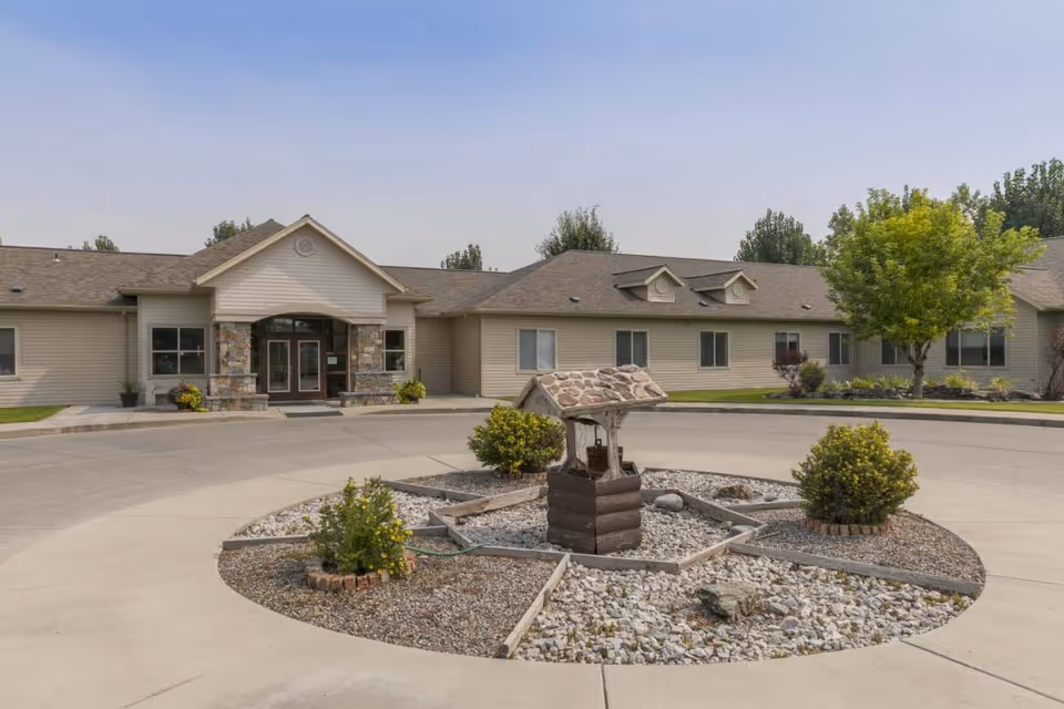 Exterior view of a single-story senior living facility with beige siding and a stone-accented entrance. The building is surrounded by a circular driveway with a landscaped center featuring a decorative wishing well and small bushes. Trees and shrubs are planted around the building under a clear sky.