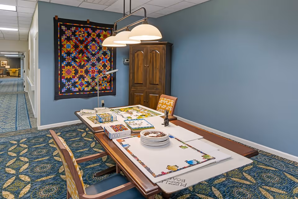 Activity room with a table set up for jigsaw puzzles, chairs, a hanging lamp, a colorful quilt on the wall, and a wooden cabinet.