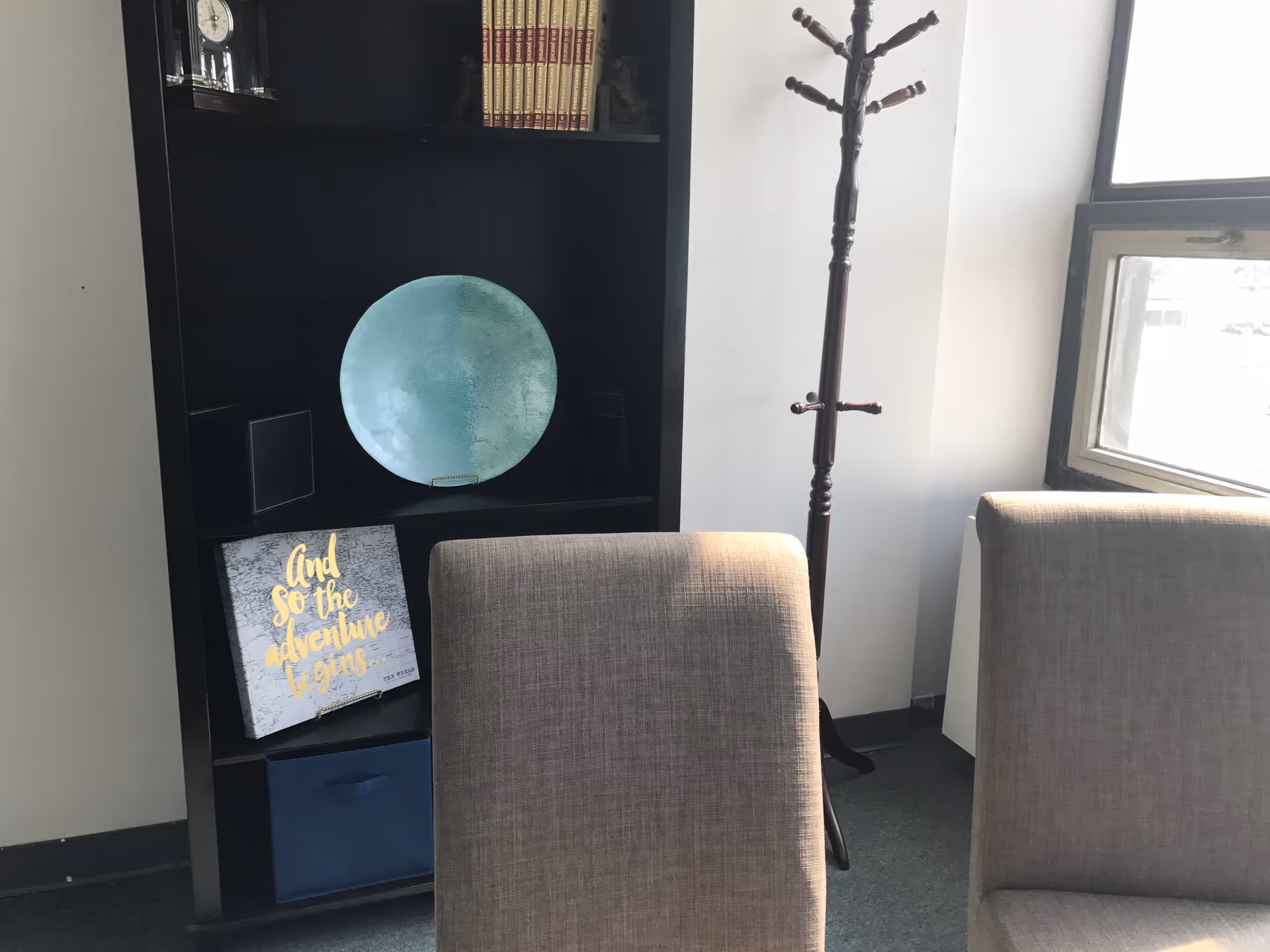 Interior living area with two beige chairs, a black bookshelf, decorative plate, coat rack, and window.