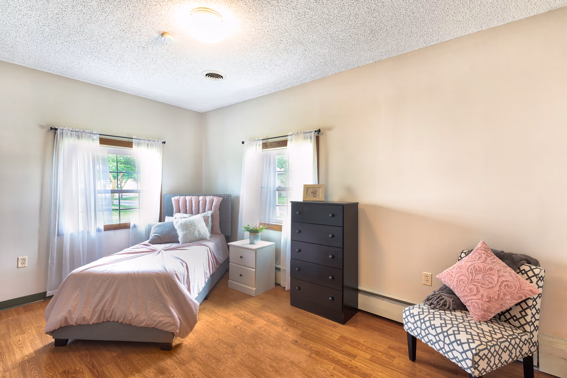 A cozy bedroom with a single bed, dresser, patterned chair, and sheer-curtained windows.