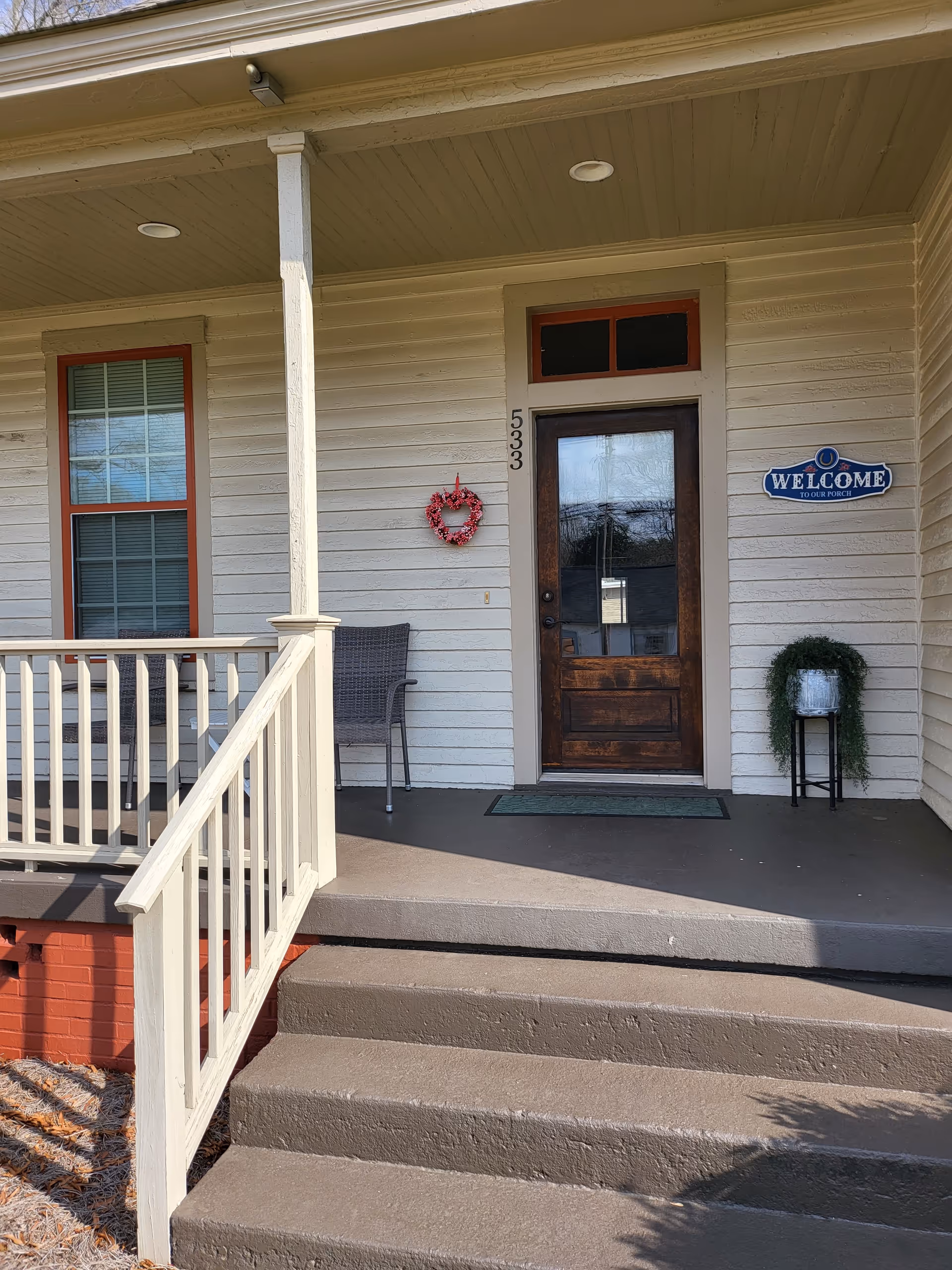 Front porch of a house with beige siding and a wooden door with glass panels. The house number 533 is displayed vertically next to the door. A small red wreath hangs on the wall beside the door. There is a single chair on the porch and a small plant on a stand near the door. A blue sign on the wall reads 'WELCOME TO OUR PORCH'. Concrete steps lead up to the porch with a white railing on the left side.