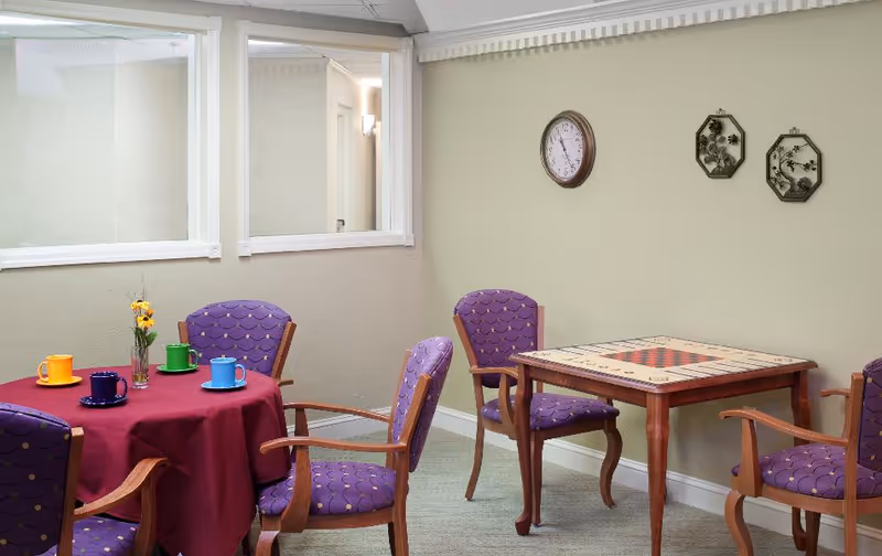 A senior living common room with purple upholstered chairs around a round table set with colorful cups and a nearby game table against a pale green wall.