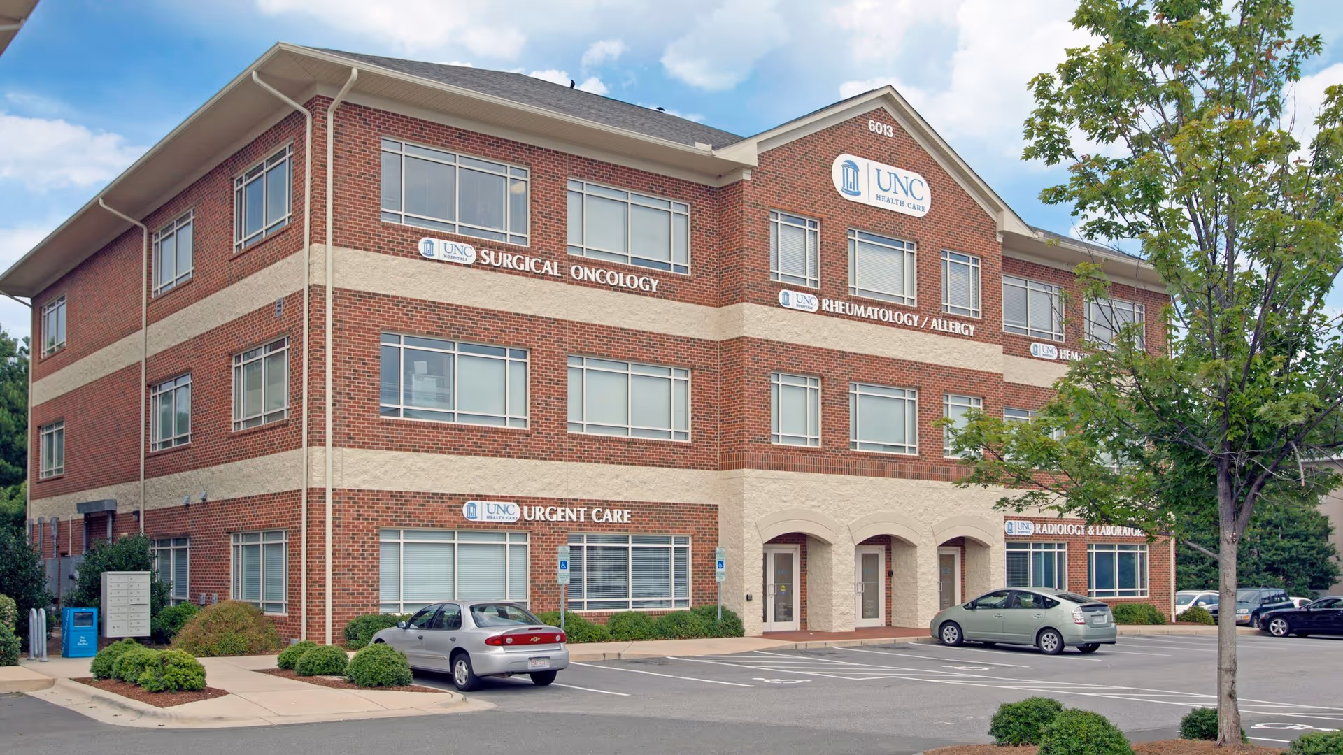 Three-story brick medical building with UNC Health Care signage and a parking lot.