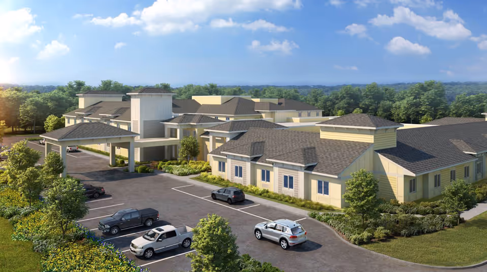 Exterior view of HearthStone at Leesburg, a large senior living facility with multiple connected buildings, a covered entrance, parking lot with several cars, surrounded by greenery and trees under a partly cloudy sky.