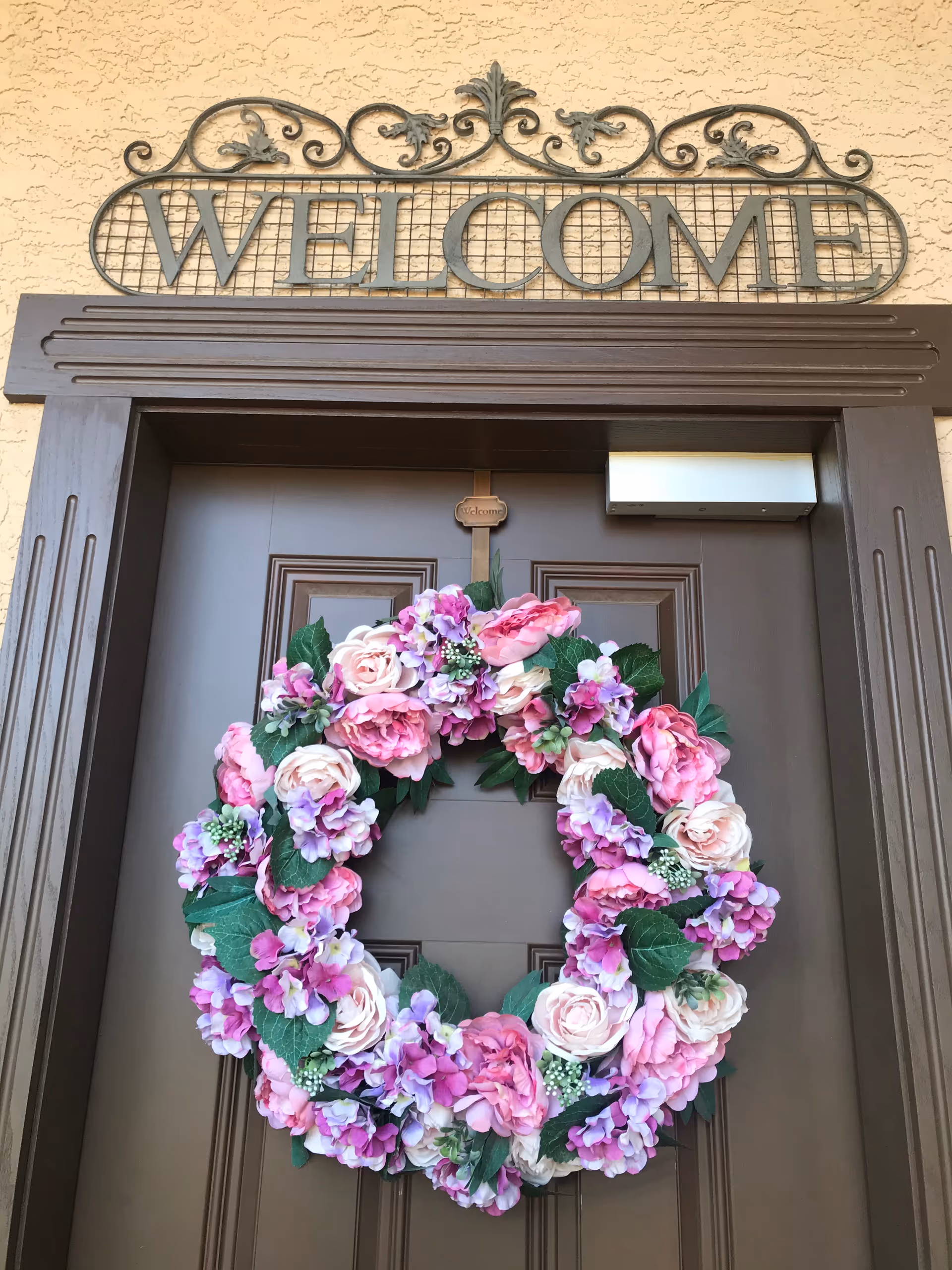 Decorative pink and purple floral wreath on a brown front door beneath a Welcome sign.