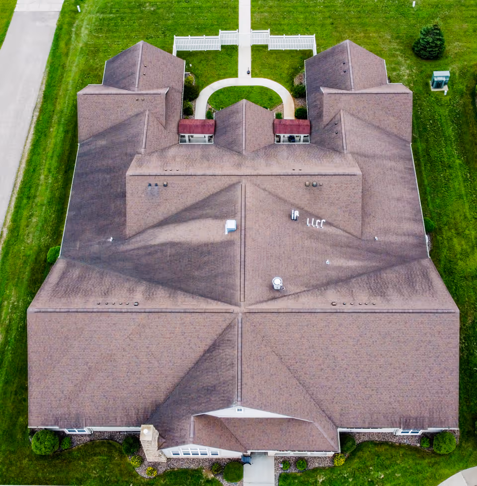 Aerial view of a large building with a brown roof surrounded by green grass and a paved walkway leading to a white fence at the top of the image.