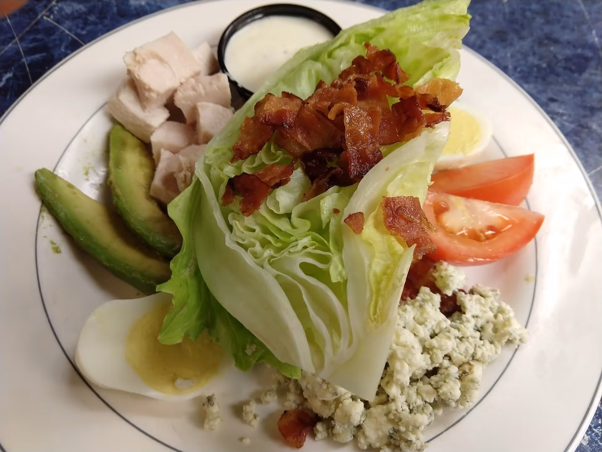 A plate of wedge salad with iceberg lettuce topped with crispy bacon bits, blue cheese crumbles, tomato wedges, sliced avocado, diced chicken, hard-boiled egg halves, and a small container of ranch dressing on the side.