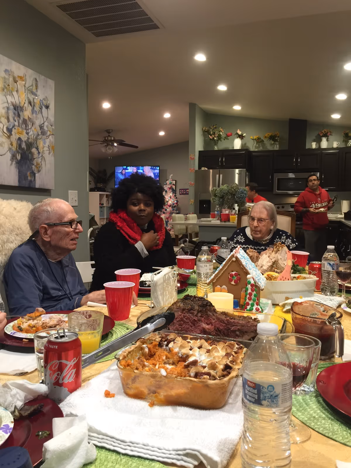 A group of people gathered around a dining table filled with various dishes including a casserole, roast meat, and a gingerbread house. The setting is a cozy kitchen and dining area with dark cabinets, a refrigerator, and a television in the background. The people appear to be enjoying a meal together in a warm, home-like environment.
