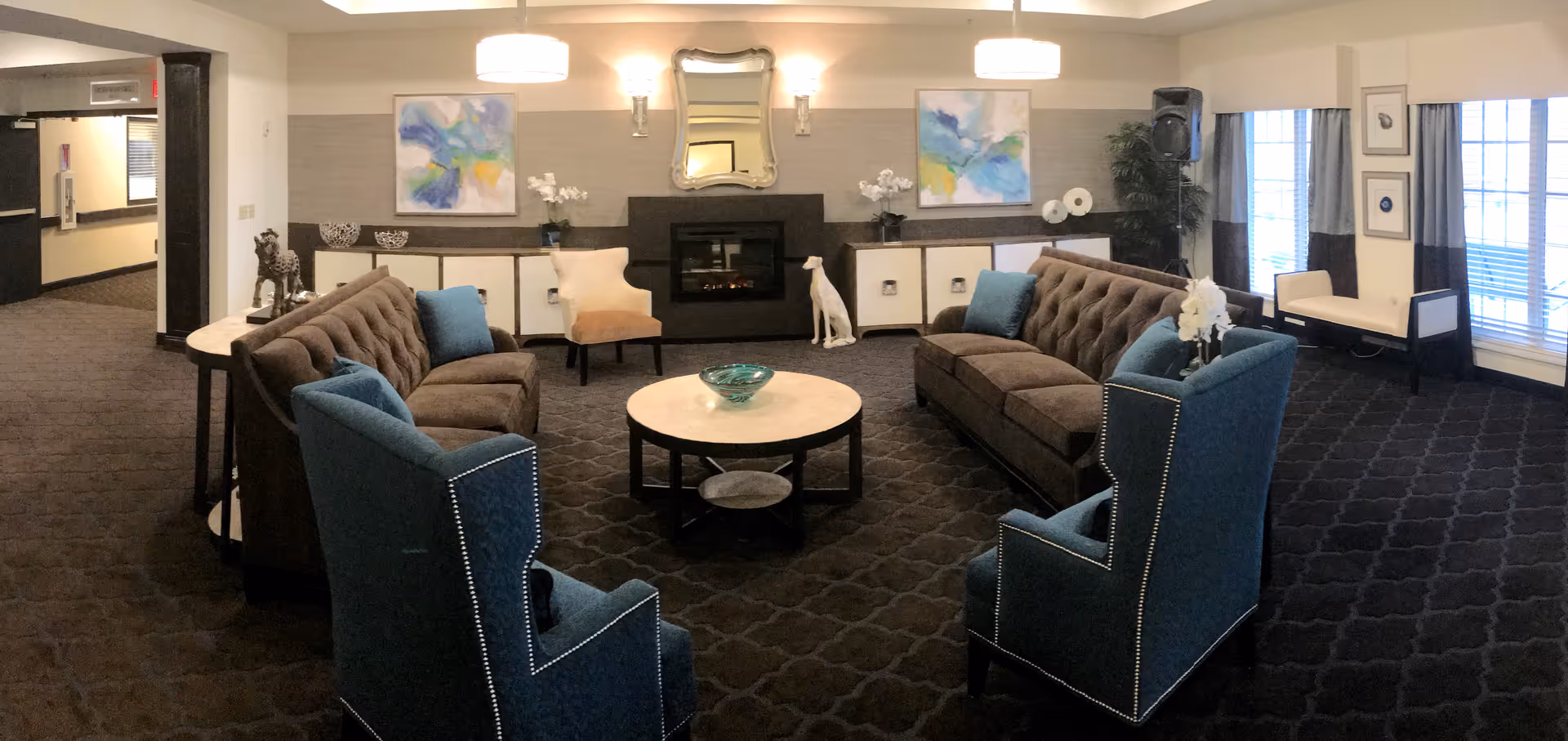 A cozy living room area with two brown sofas, two blue armchairs, and a round coffee table in the center. The room features a fireplace with a decorative mirror above it, two abstract paintings on the wall, and a white dog statue next to the fireplace. There are large windows with curtains on the right side and a hallway visible on the left.