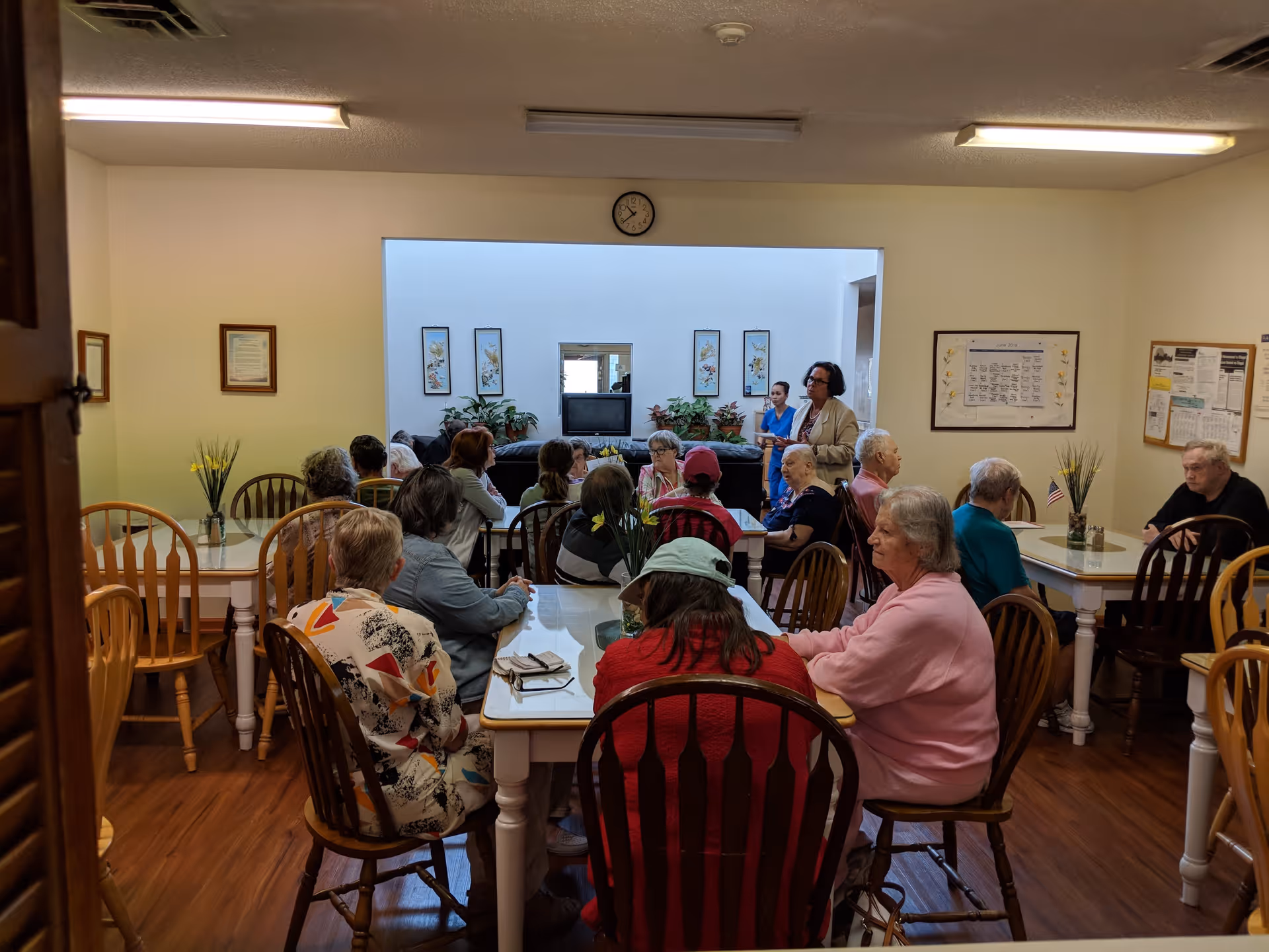 A group of elderly people seated around tables in a communal room, with two staff members standing and interacting with them. The room has wooden chairs, framed pictures on the walls, and a clock above an open window-like space showing another room with plants and a TV.