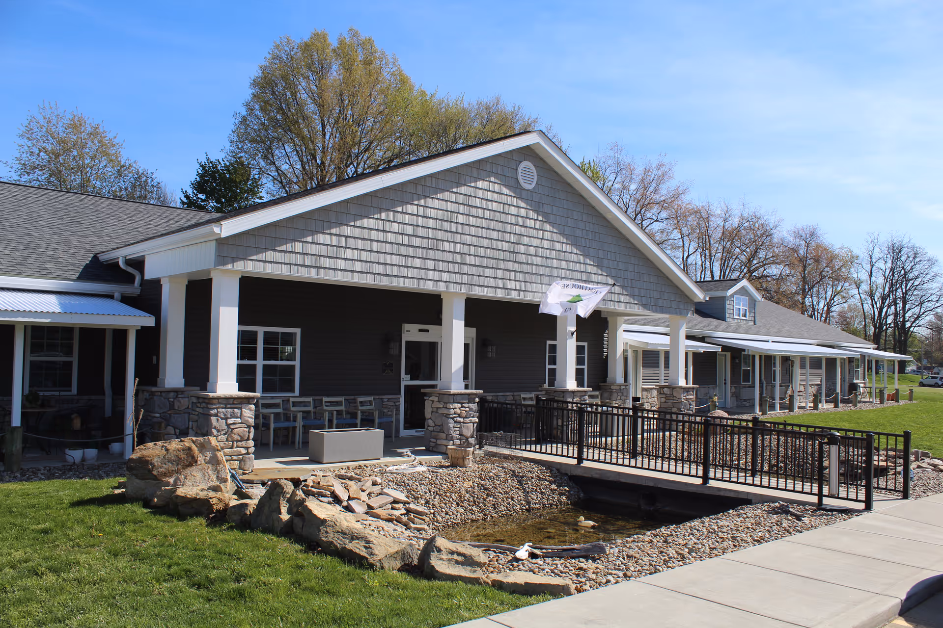 Exterior view of a single-story senior living facility building with a covered porch supported by white columns and stone bases. There is a small pond with rocks and a black metal railing bridge in front of the entrance. Trees and a clear blue sky are visible in the background.