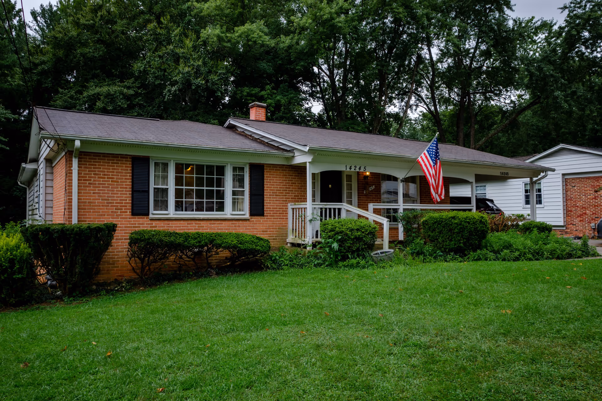 Front exterior view of a single-story brick house with a small porch, an American flag mounted near the entrance, surrounded by green bushes and a well-maintained lawn, with tall trees in the background.