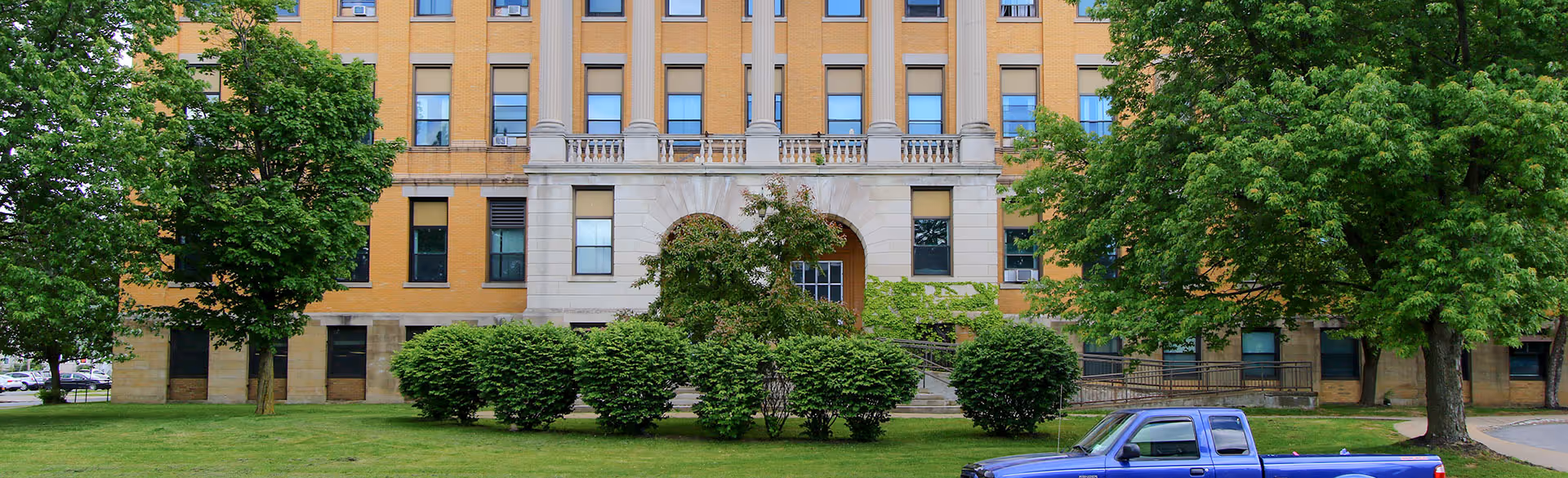 Front of Focus Senior Living at Utica building with trees, shrubs, and a blue pickup.
