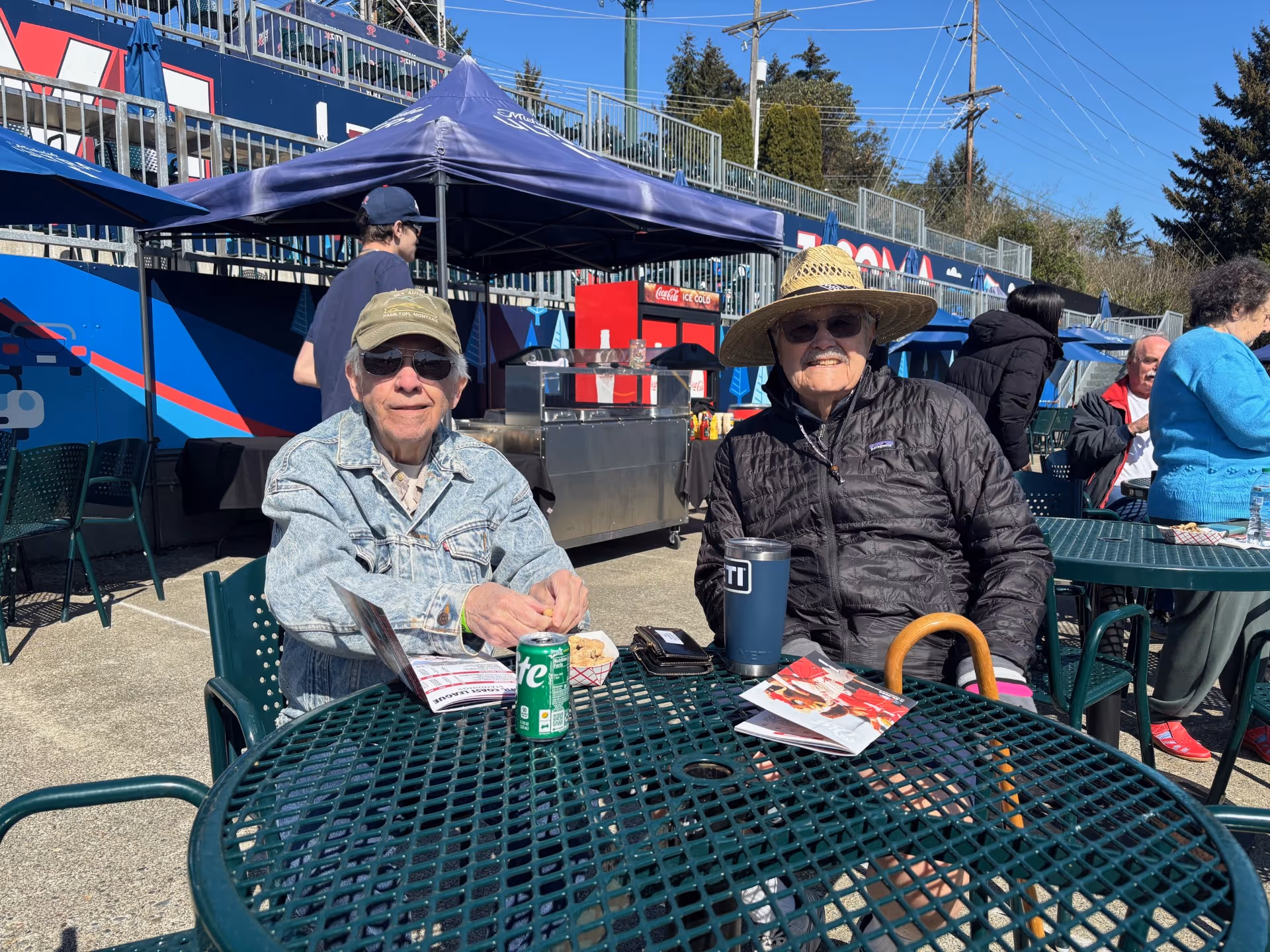 Two elderly men sitting at a green metal outdoor table under a clear blue sky. One man is wearing a denim jacket, sunglasses, and a cap, while the other is wearing a black jacket, sunglasses, and a straw hat. There are other people sitting at nearby tables and a concession stand with a blue canopy in the background.