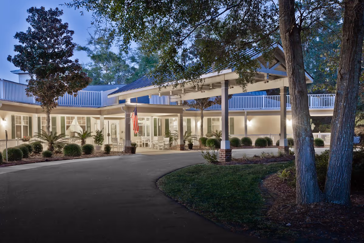 Covered entrance and driveway of a senior living building at dusk with an American flag, porch seating, and surrounding trees.