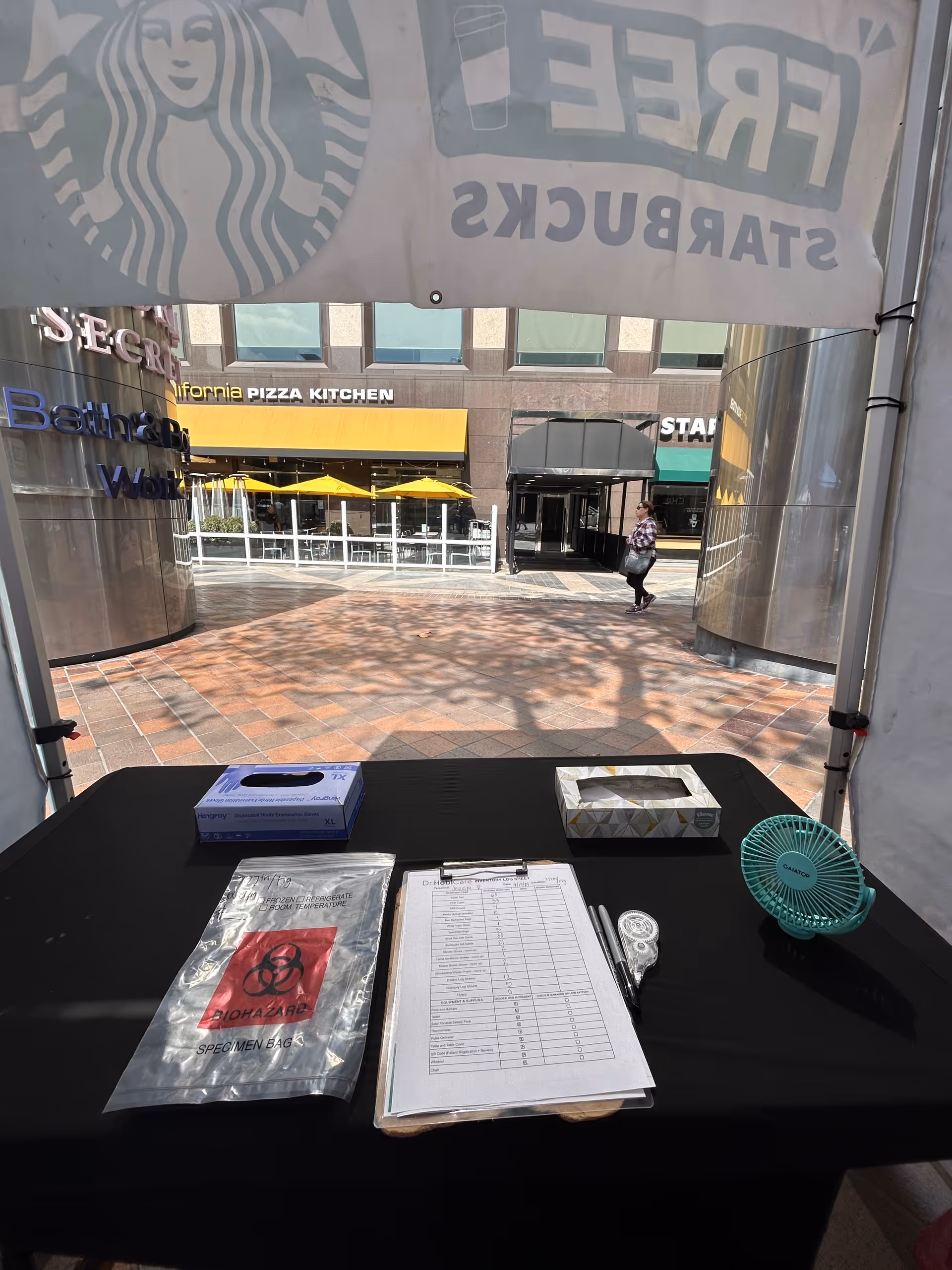 Outdoor tent setup with paperwork, a biohazard bag, and a table in front of mall storefronts.