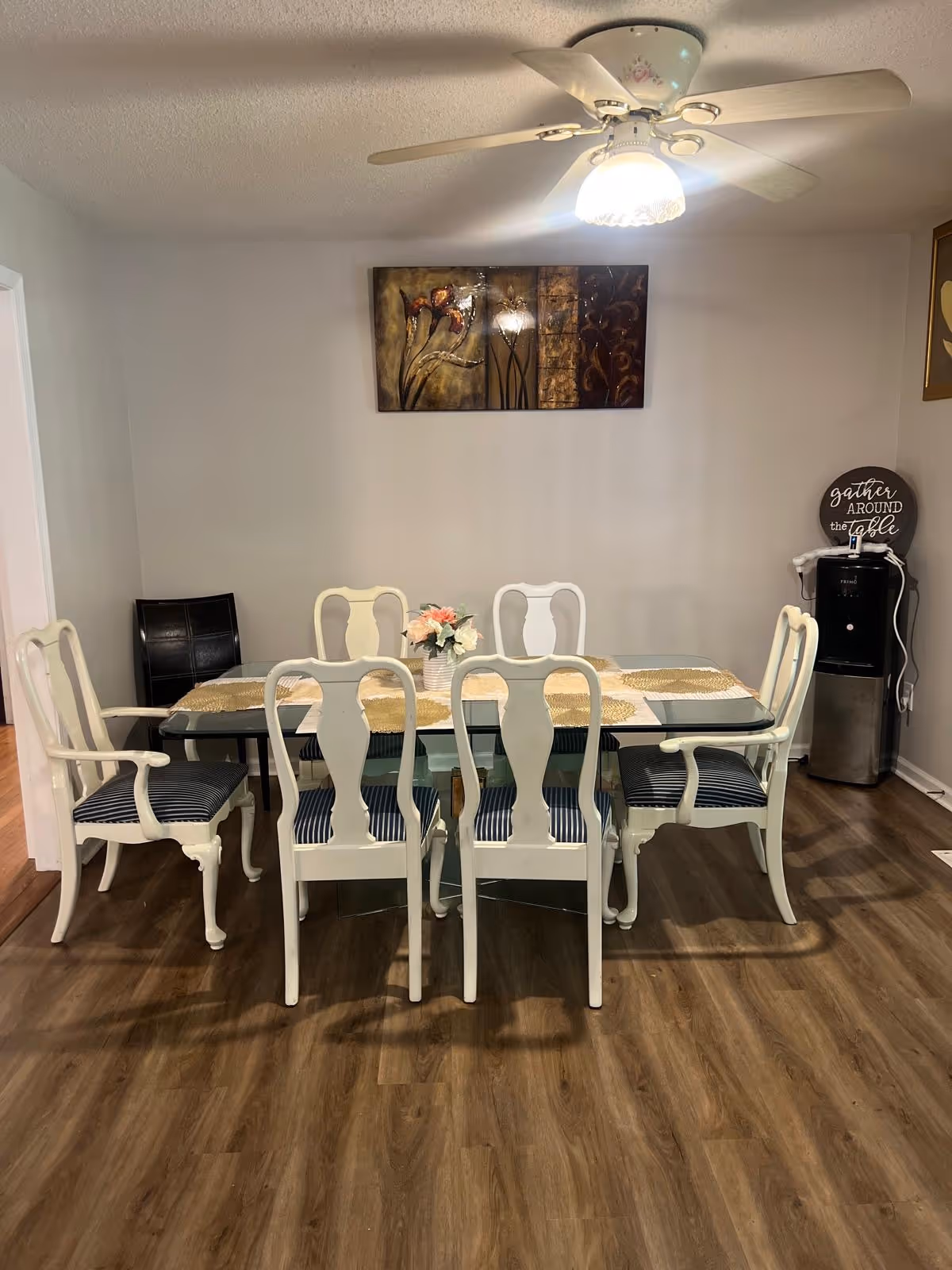 Dining room with glass table, eight white chairs, wall art, and a ceiling fan.