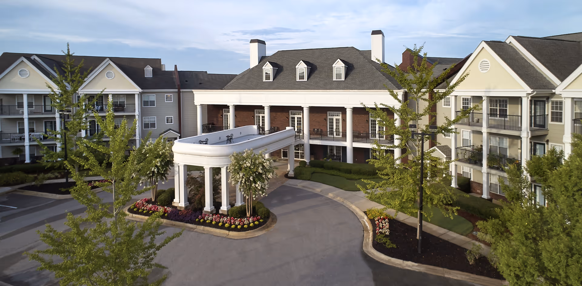Front exterior view of Redstone Village senior living facility showing a large building with white columns, multiple windows, balconies, and a covered driveway entrance surrounded by landscaped trees and flower beds.
