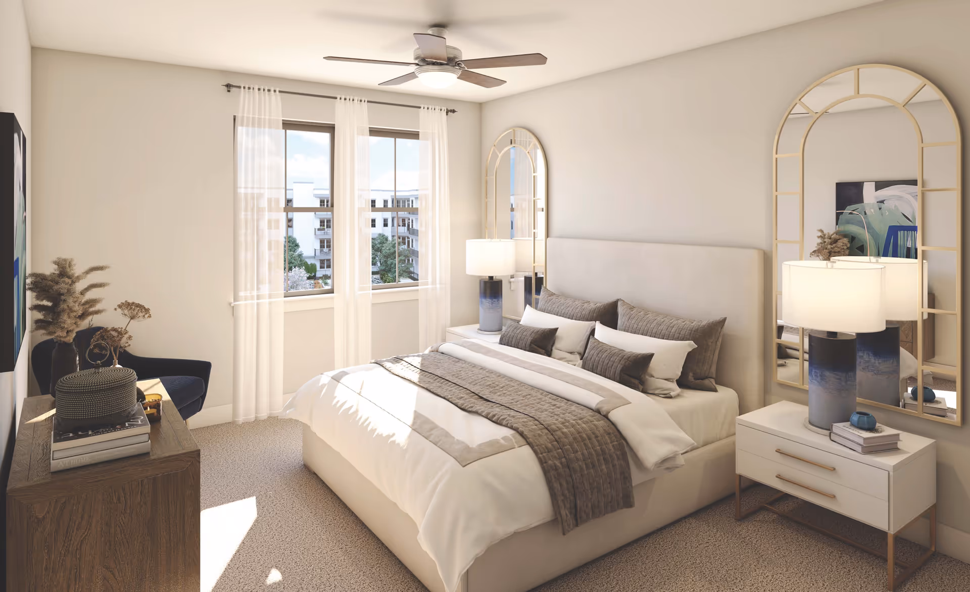 A bright and cozy bedroom with a large bed featuring white and beige bedding and multiple pillows. There are two tall arched mirrors behind the nightstands on either side of the bed, each with a blue and white lamp. A ceiling fan with a light is mounted on the ceiling. A window with sheer white curtains lets in natural light, showing a view of a building and trees outside. A wooden dresser with books and decorative items is positioned near a dark blue armchair.
