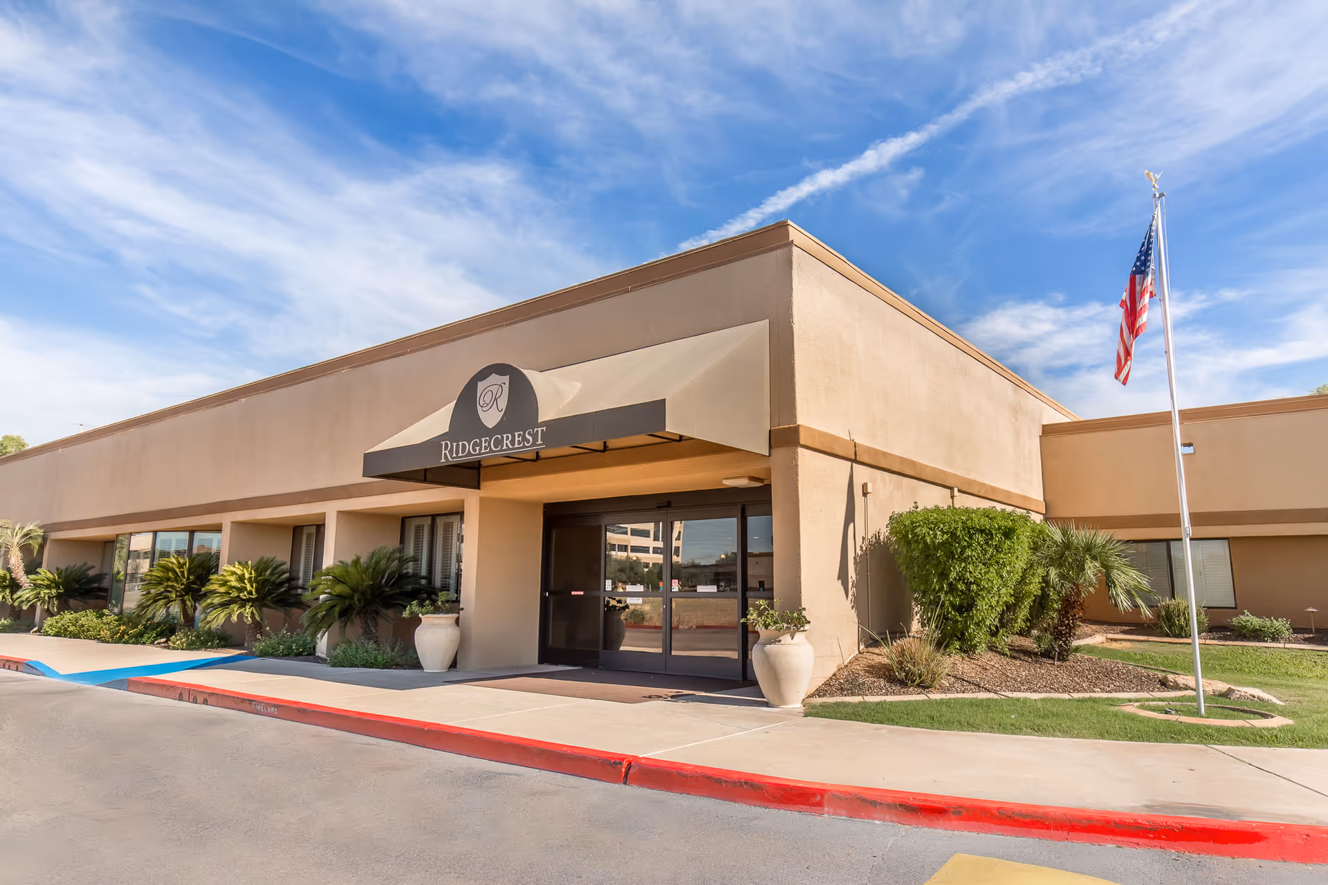 Exterior view of Ridgecrest Post Acute facility showing the entrance with a beige awning displaying the Ridgecrest logo and name. The building is single-story with tan walls, surrounded by landscaped greenery and an American flag on a flagpole to the right.