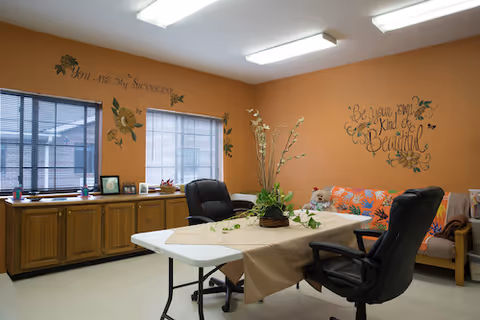 Orange-walled common room with a table, chairs, windows, and a sofa with decorative pillows.
