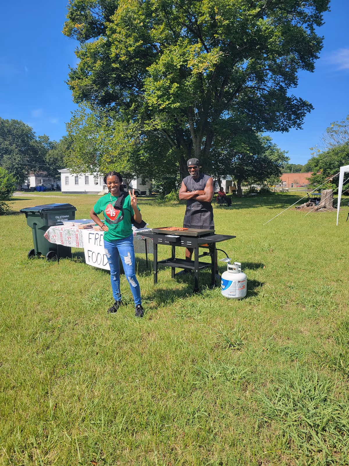 Two people stand near a grill on a grassy lawn with a table offering free food.