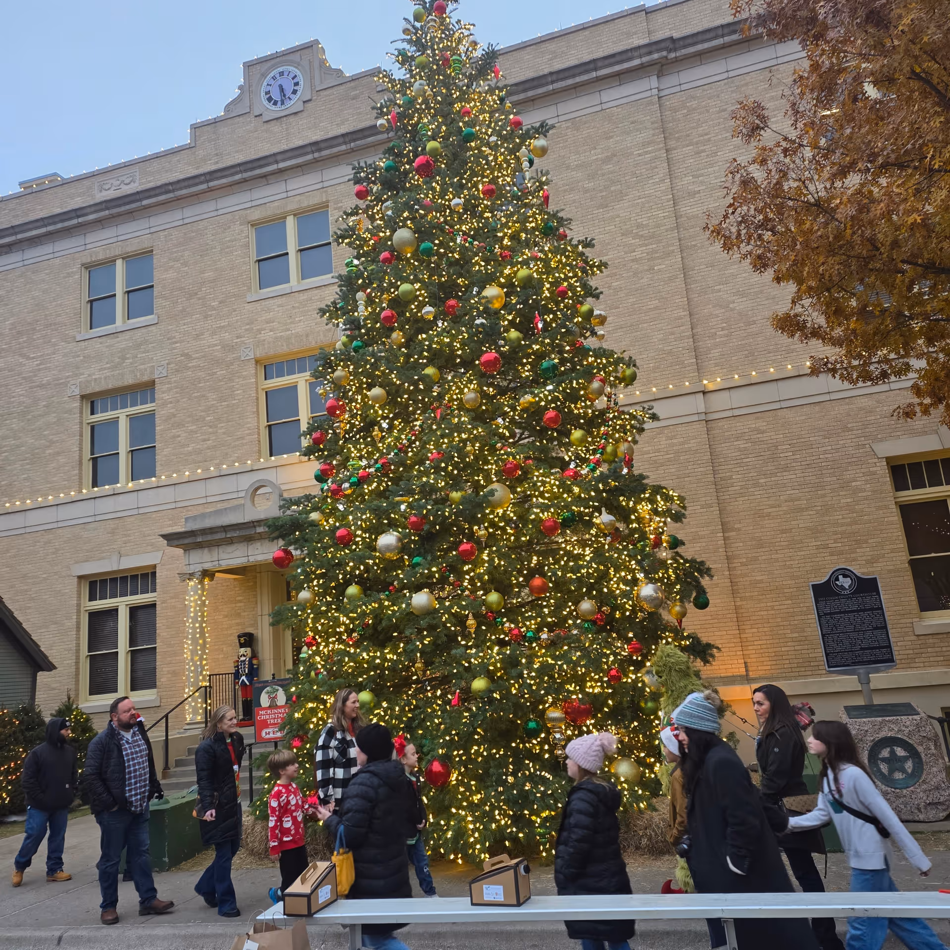 A tall, ornamented Christmas tree illuminated with lights outside a brick building, surrounded by people.