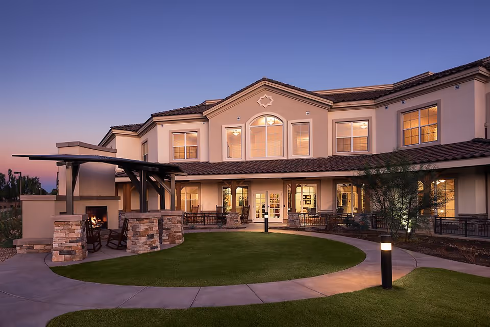 Two-story residential building at dusk with lit windows, a circular lawn, walkway, and covered outdoor seating area with a fireplace.