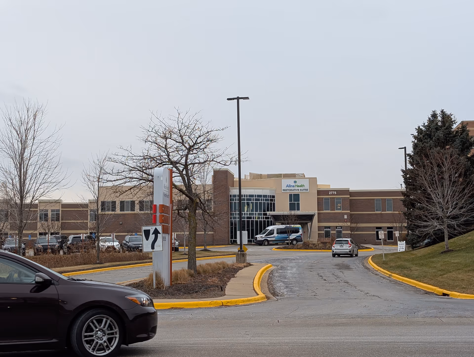 Front entrance of a low-rise healthcare building with a circular drive, parked cars, and bare trees on an overcast day.