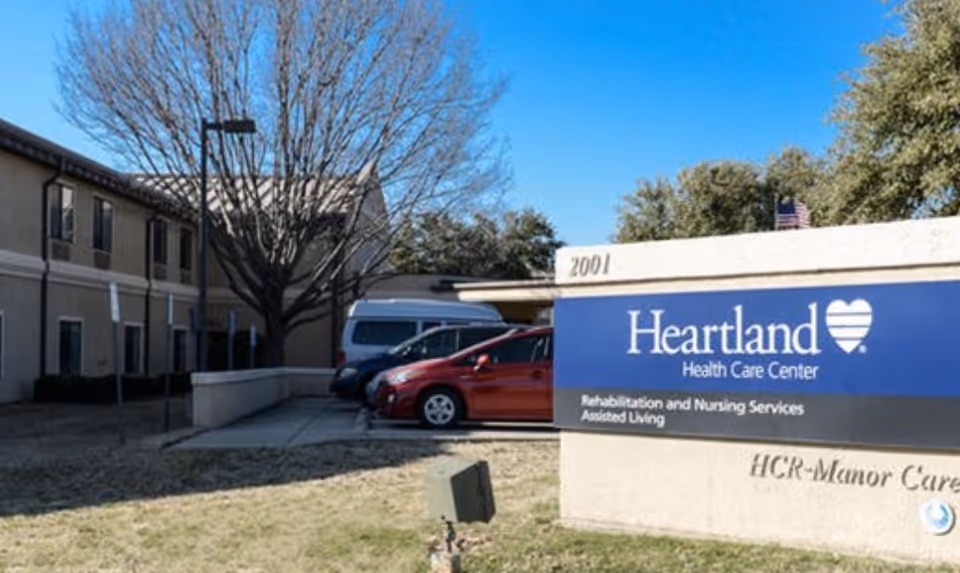 Front exterior of Heartland Health Care Center with a large blue sign, parked cars, and the two-story building behind.