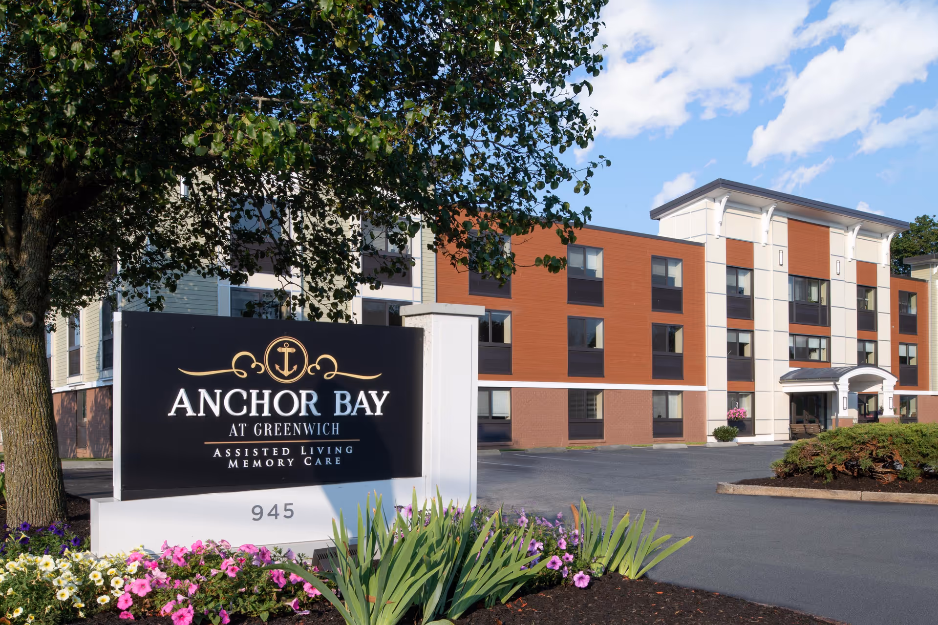 Front exterior of Anchor Bay at Greenwich assisted living facility with a prominent entrance sign and landscaped flowerbeds.