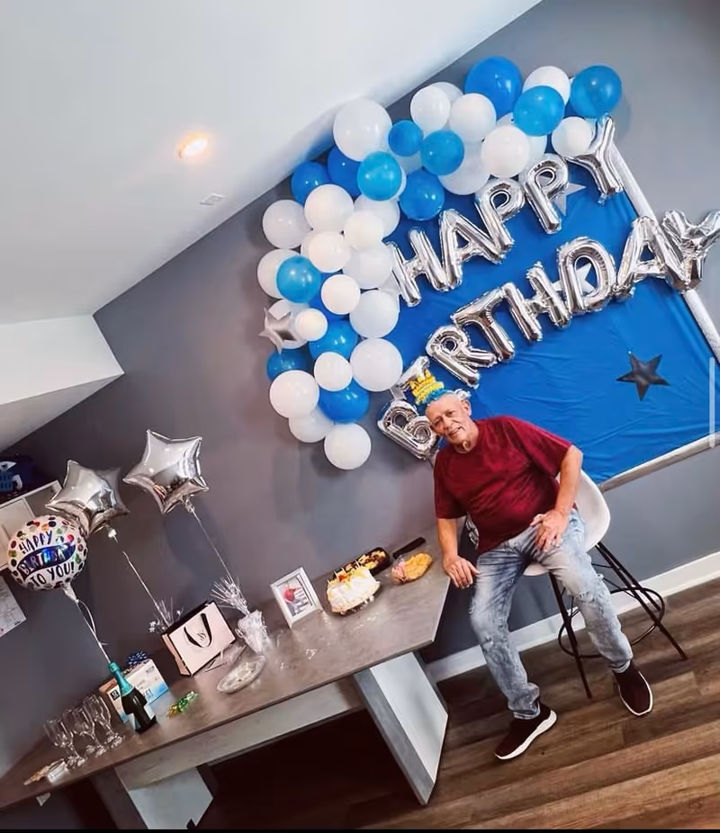 Indoor birthday party setup with blue and white balloons and a man beside cake.