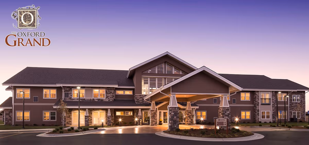 Exterior view of The Oxford Grand Assisted Living & Memory Care building at dusk with lights glowing warmly from the windows and a covered entrance in front.