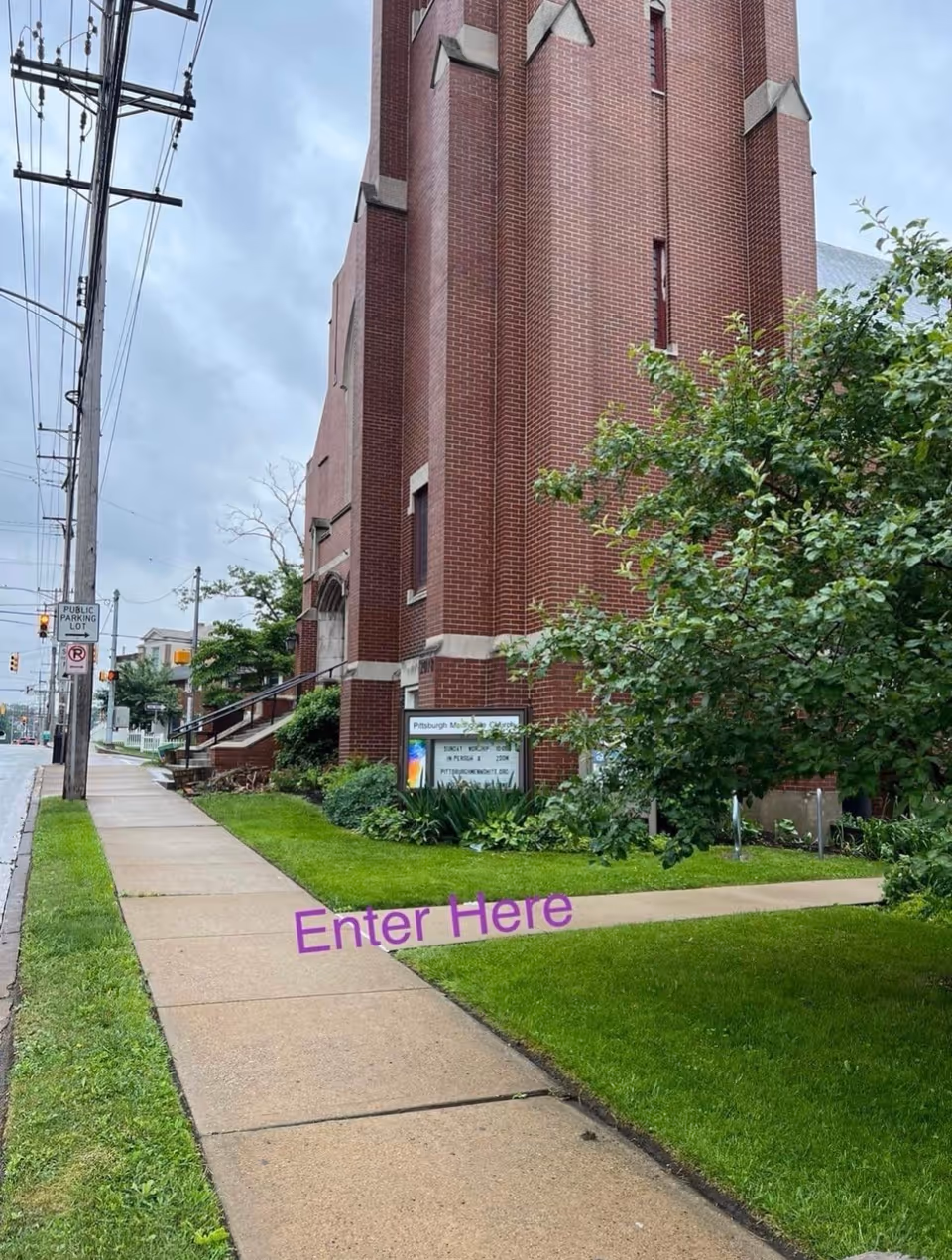 Tall red brick building beside a sidewalk with green lawn, trees, and an Enter Here sign.