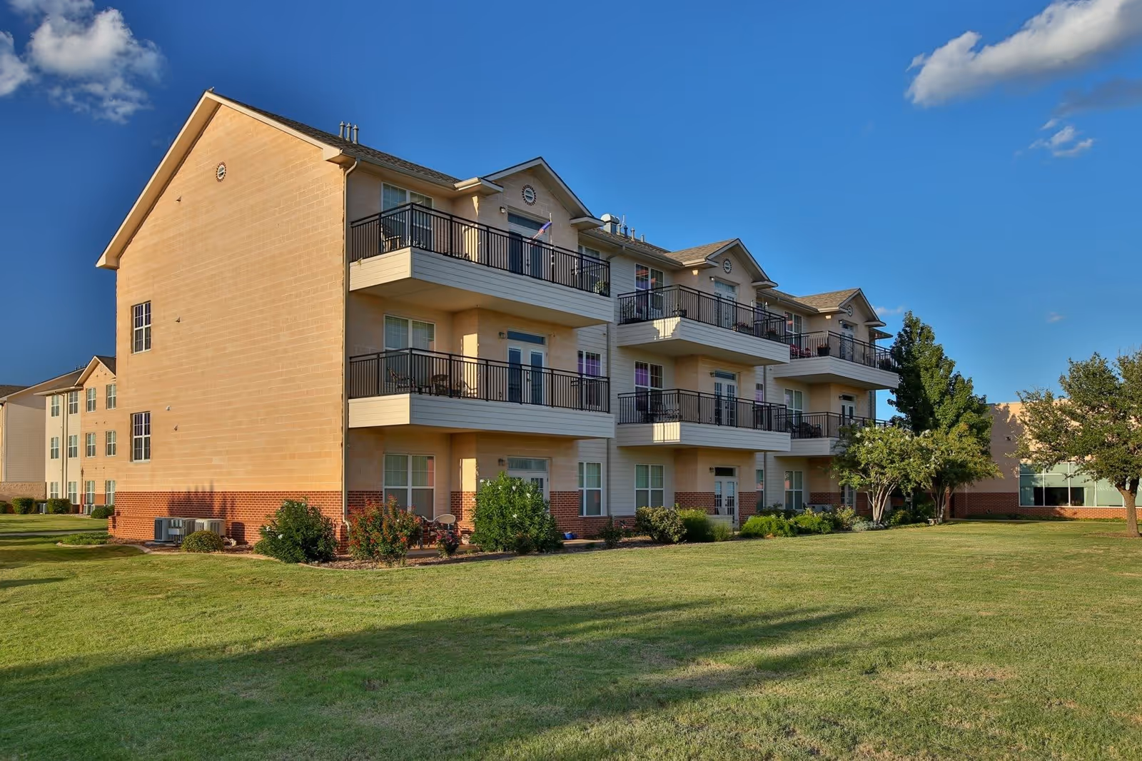 Exterior view of a three-story residential building with balconies on each floor, surrounded by a well-maintained lawn and some trees under a clear blue sky.