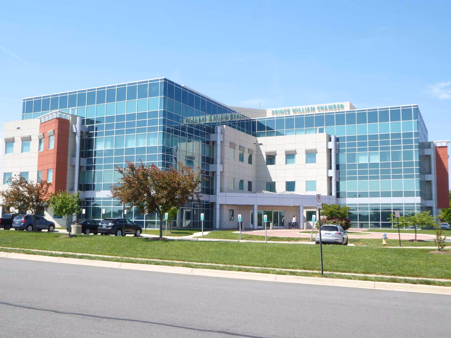 Modern glass-front building with a main entrance, green lawn, and parked cars.