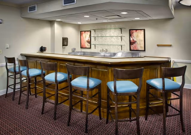 A wooden bar counter with blue-cushioned stools inside a senior living facility lounge.