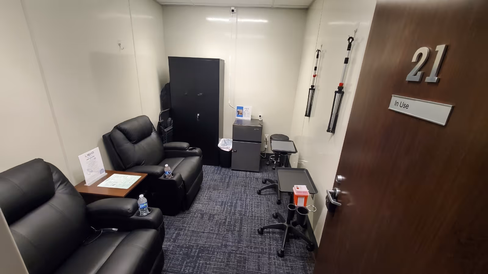 A small interior treatment room with two black recliners, IV poles, and medical equipment.