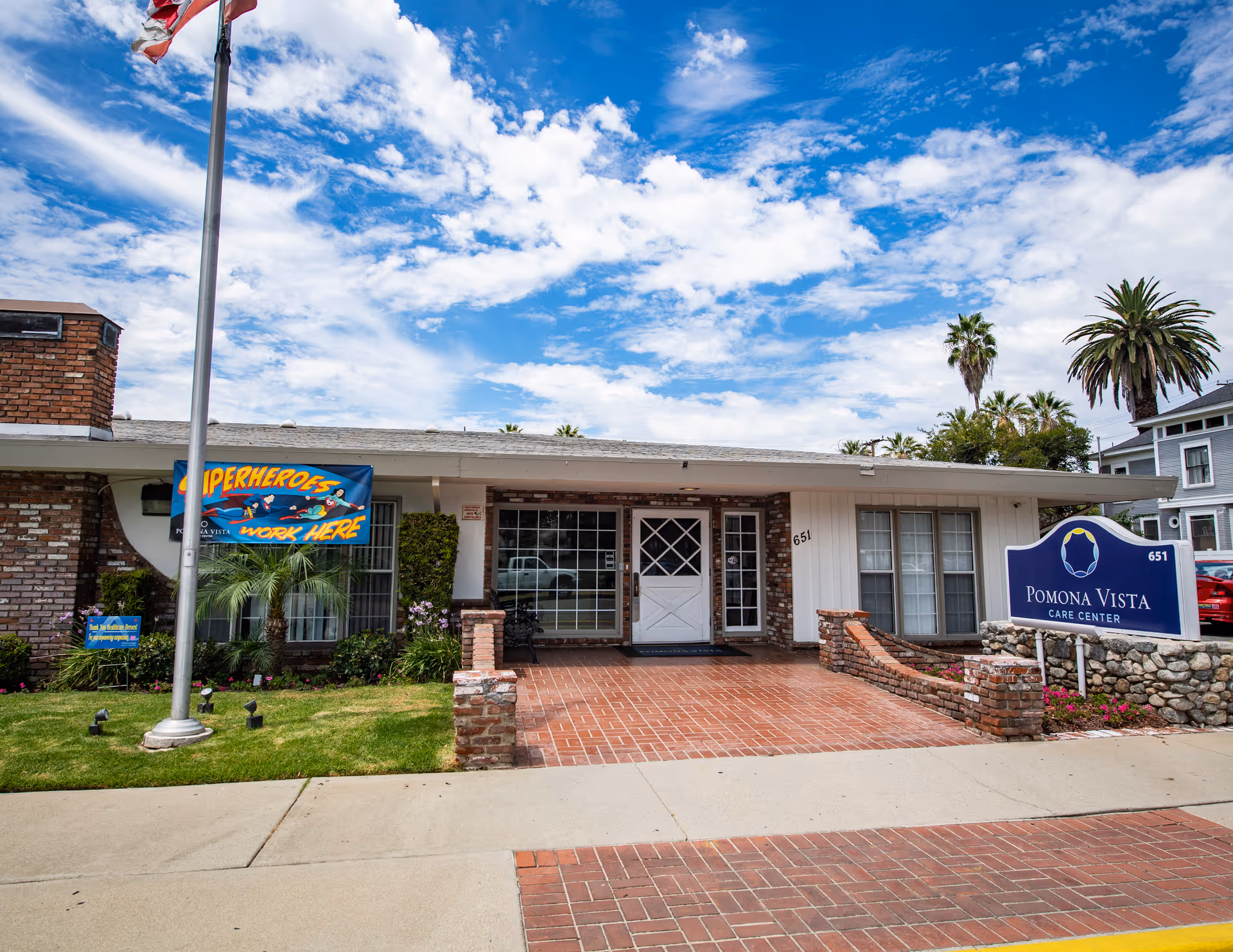 Exterior view of Pomona Vista Care Center building with a brick and white facade, a flagpole with an American flag, a blue sign displaying the facility name, and a banner that reads 'Superheroes Work Here'. The sky is partly cloudy with palm trees visible in the background.