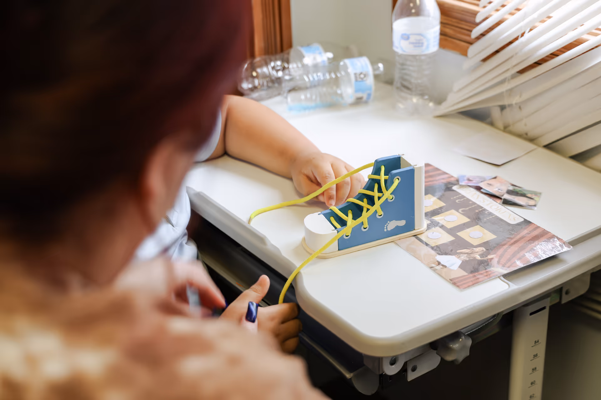 Close-up of a person tying yellow shoelaces on a blue toy shoe at a therapy desk.