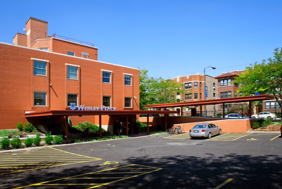 Red-brick building front with a covered entrance reading 'Wesley Place', a parking lot and a car beneath a clear blue sky.