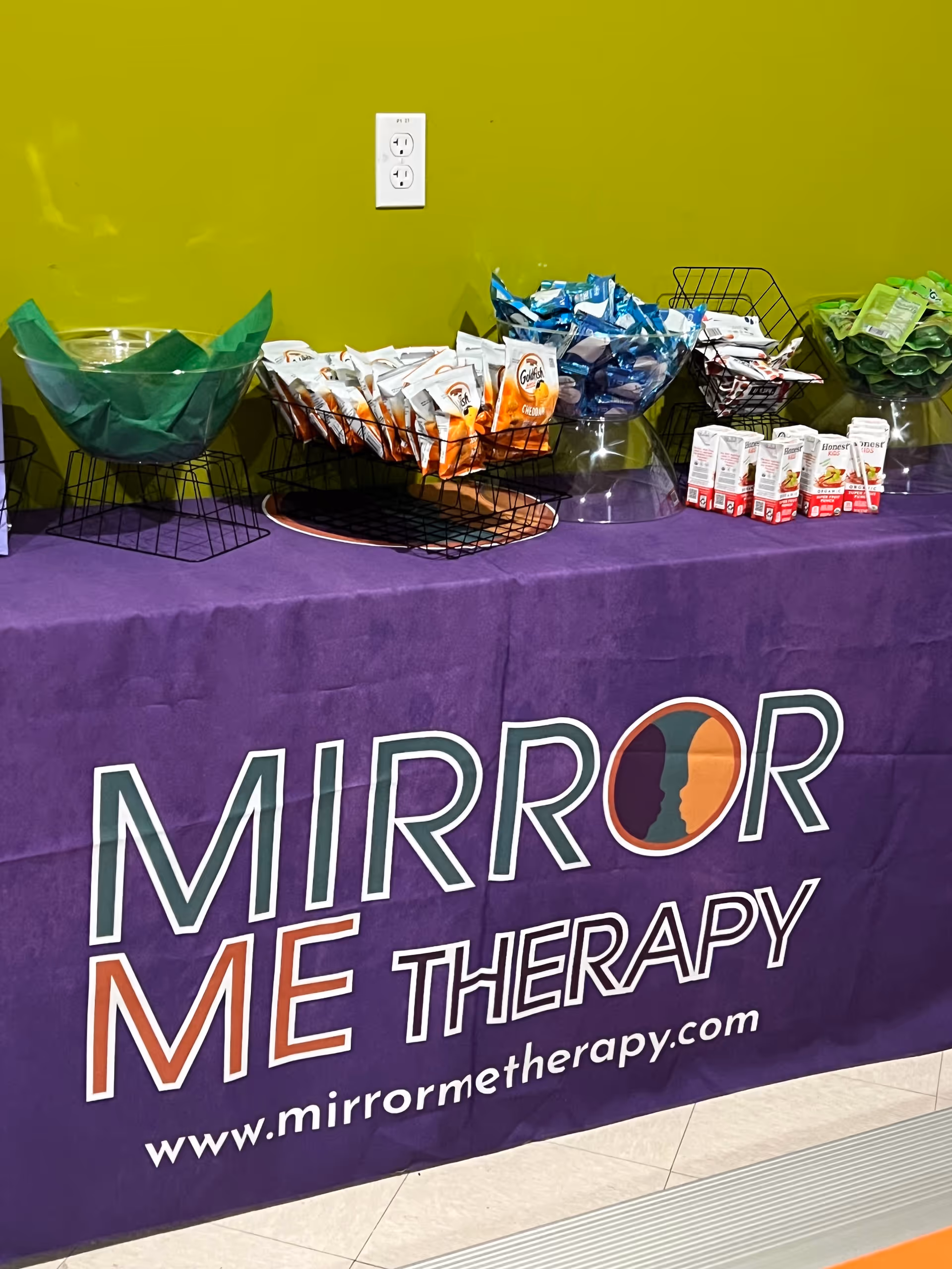 An interior display table with colorful snack baskets promoting Mirror Me Therapy against a lime wall.