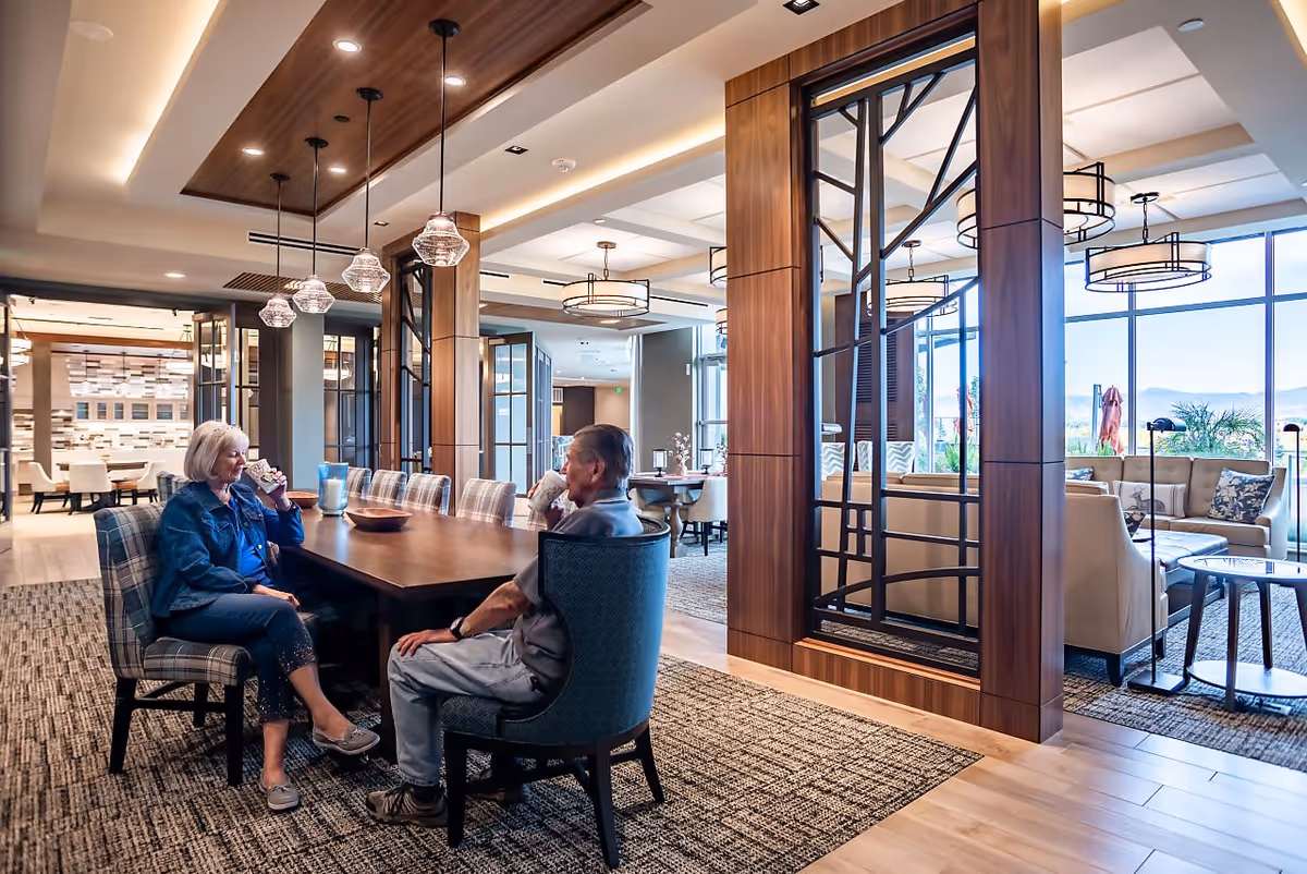 Two elderly people sitting at a long wooden table in a well-lit, modern common area with large windows, comfortable seating, and decorative lighting fixtures.
