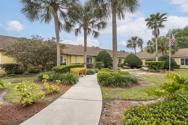 Curved walkway through a landscaped outdoor area toward a single-story yellow building with palm trees.