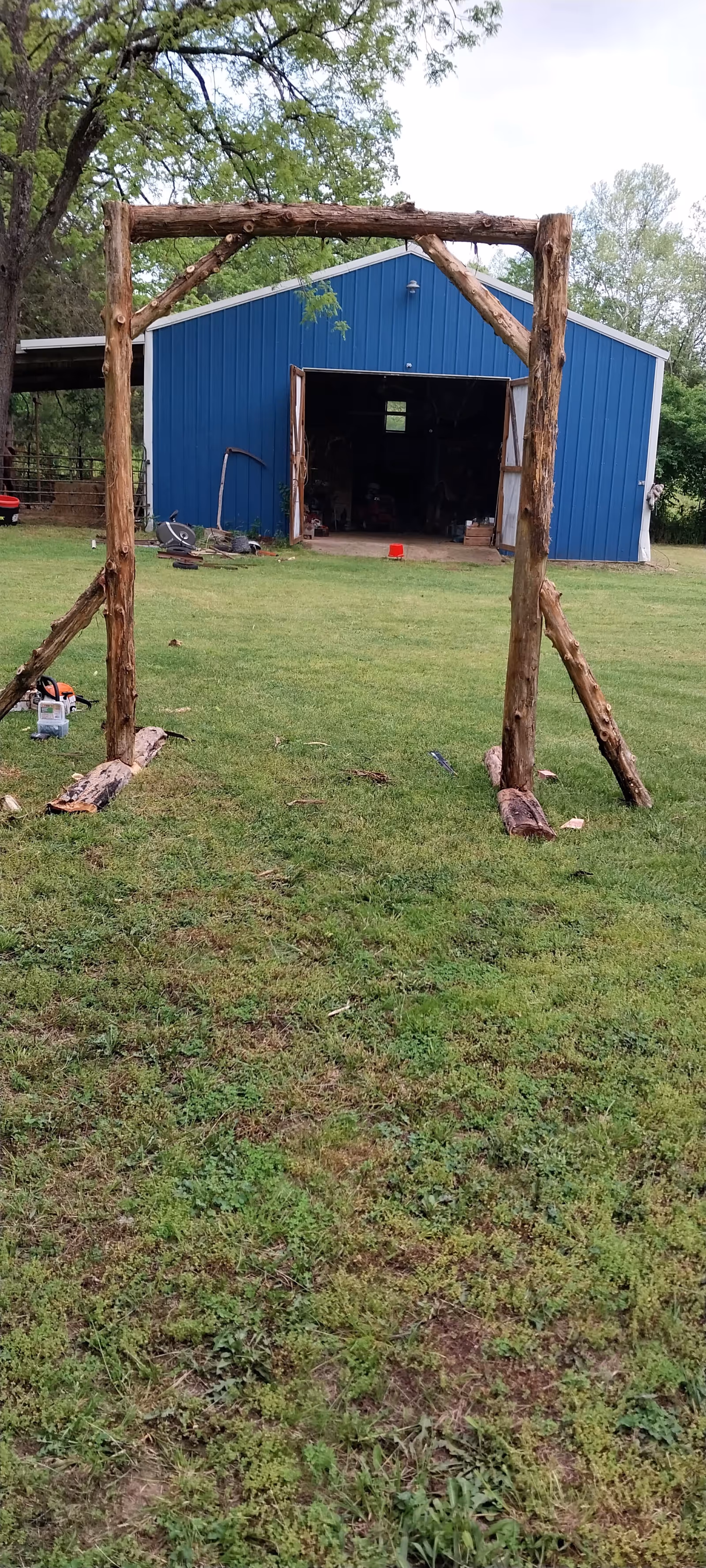 Blue metal building with a wooden arch frame in a grassy yard.