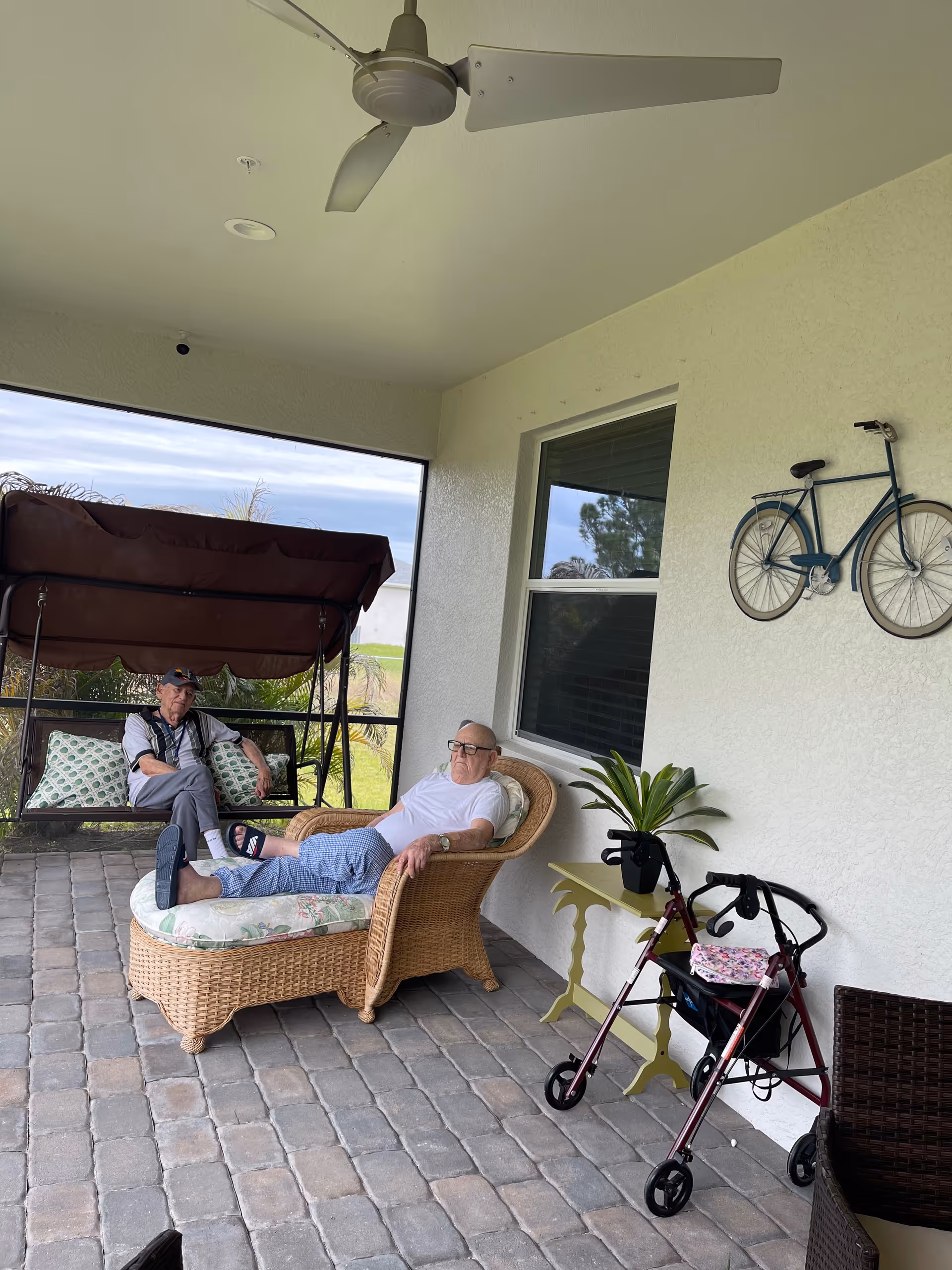 Two elderly men relax on a covered outdoor patio with wicker furniture.