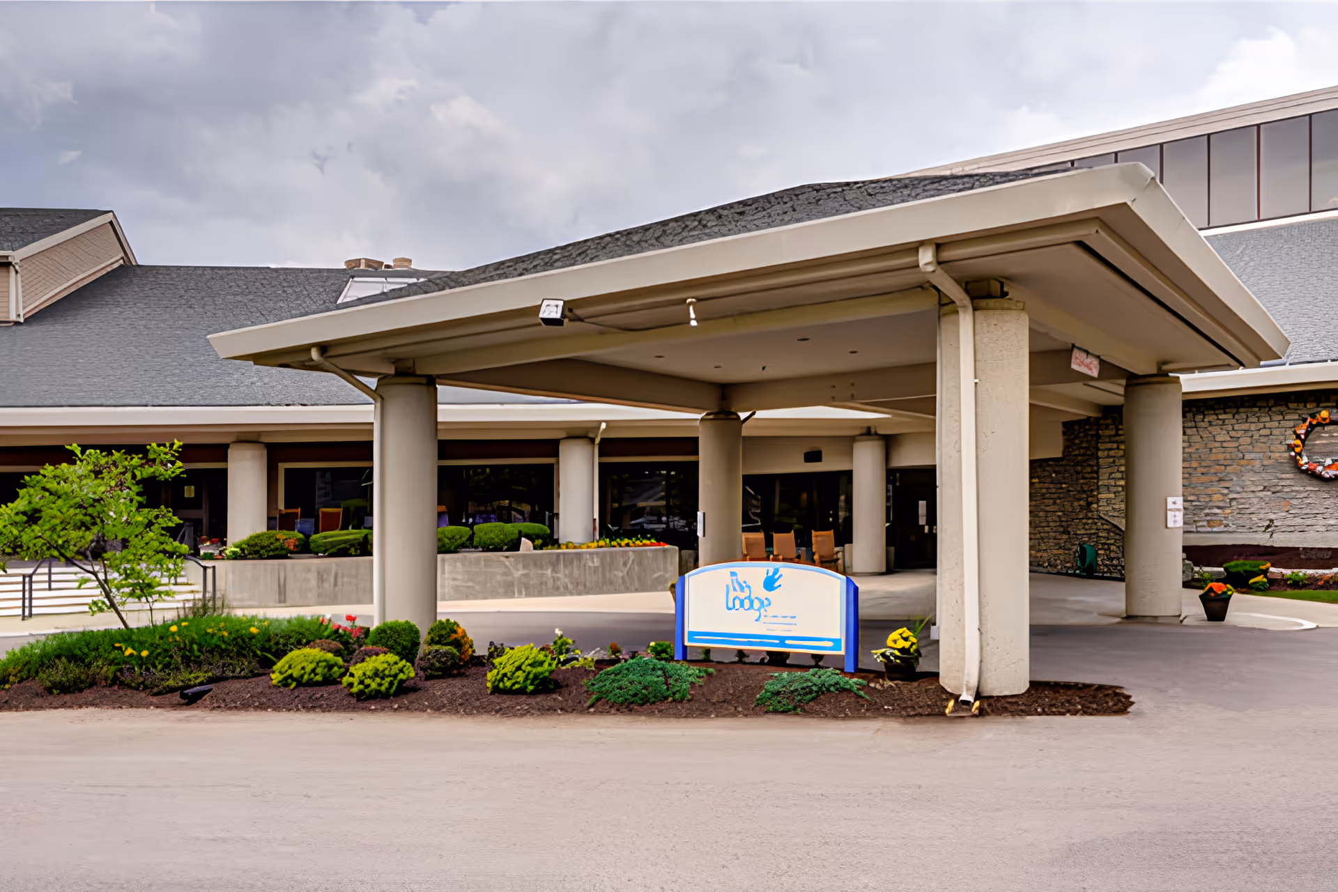 Entrance area of The Lodge Retirement Community featuring a covered drop-off zone supported by large columns, landscaped greenery, and a sign with the community's logo. The building exterior includes stone and large windows, with a cloudy sky overhead.