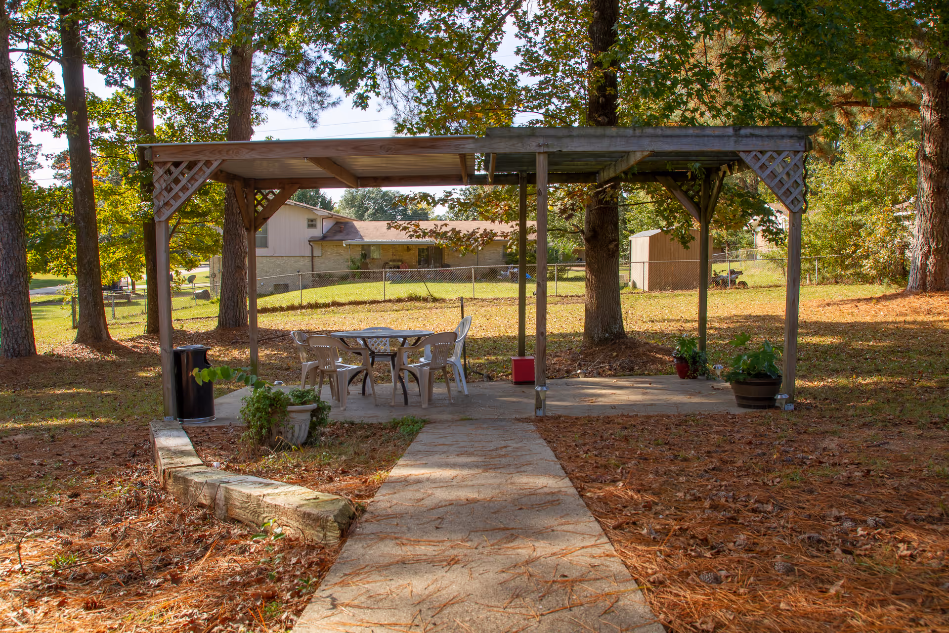 Outdoor seating area with a wooden pergola covering a round table and four plastic chairs, surrounded by trees and plants in a grassy yard with a concrete pathway leading to the seating area.