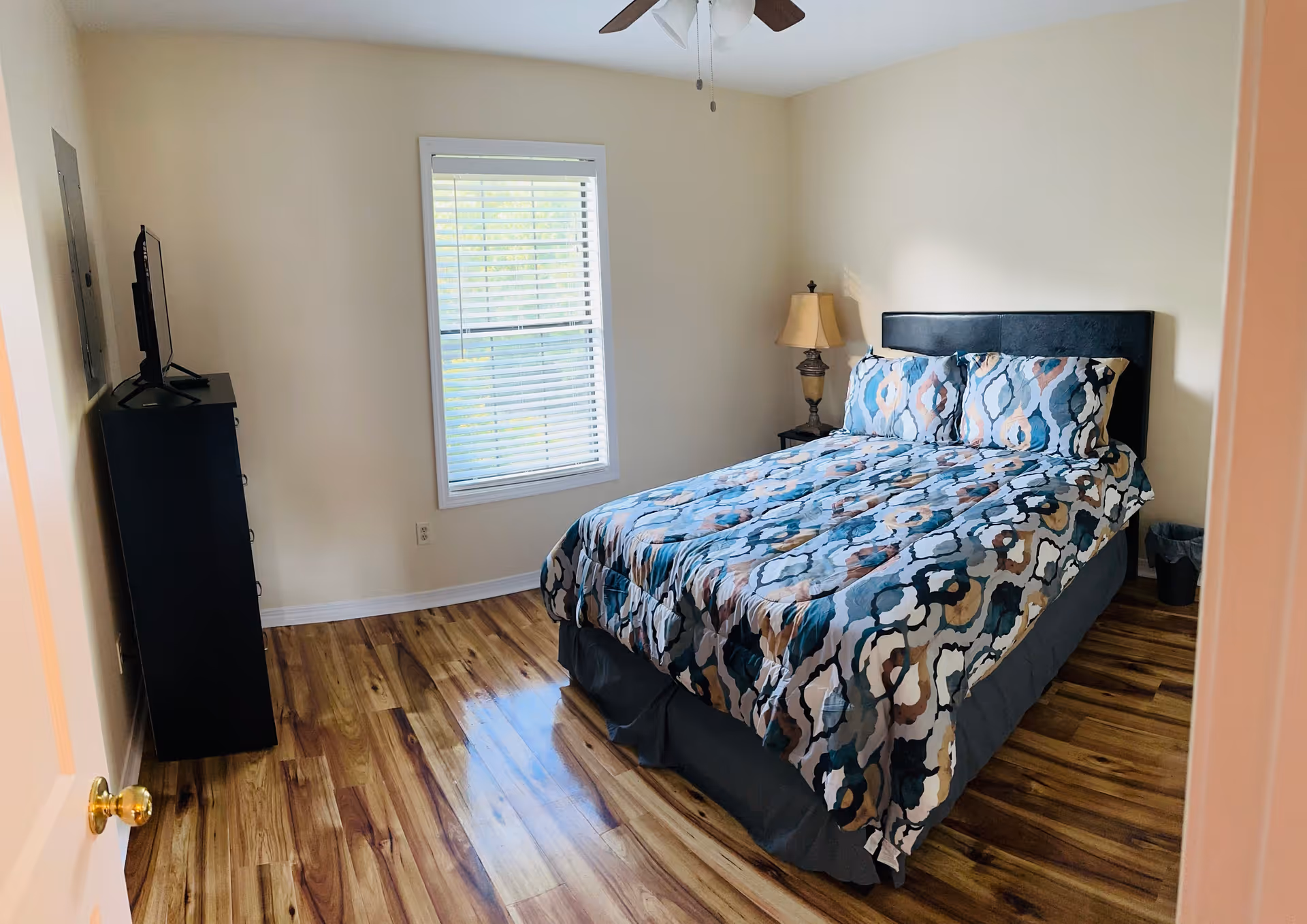 Bright bedroom with patterned bedding, nightstand lamp, and dresser.
