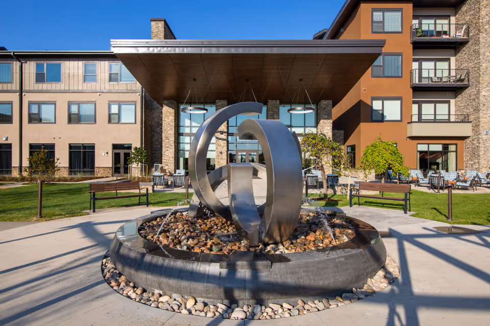 Entrance of a modern senior living building featuring a circular metal sculpture fountain, benches, and patio seating.
